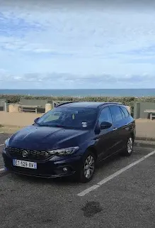 Voiture break bleu foncé stationnée face à l'océan sur un parking côtier.