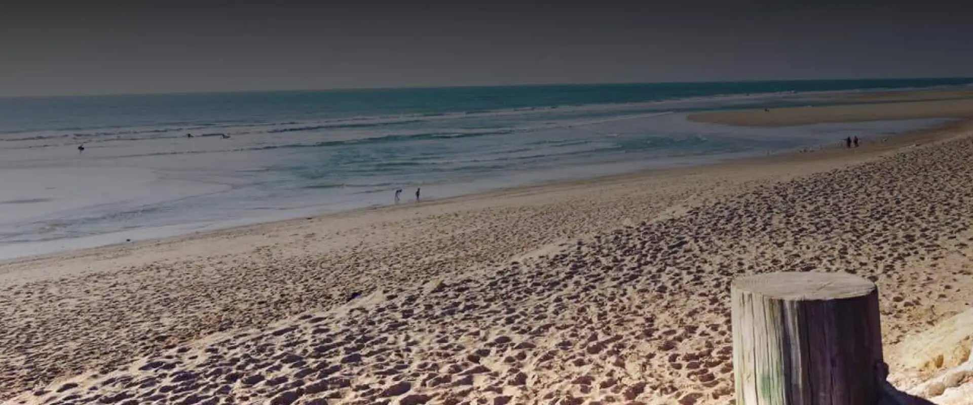 Plage de sable en bord d’océan avec des promeneurs et un pieu en bois.